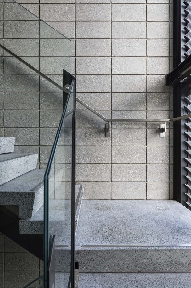Close-up architectural view of a modern stairway and landing. The space features exposed grey concrete block walls, polished grey terrazzo or concrete stairs and flooring, dark metal handrails, and clear glass balustrades. A louvred window or screen is visible along the right side, bringing in filtered light. The composition is stark, geometric, and industrial in character.