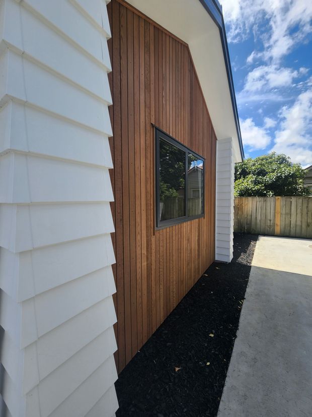 Exterior view of a contemporary residential addition or facade, featuring warm vertical timber cladding, crisp white horizontal weatherboard cladding, a dark-framed window, and a simple roof overhang. A concrete driveway and fenced side yard are visible under bright daylight.
