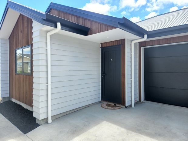 Exterior view of a contemporary single-storey home entry and attached garage. The facade combines white horizontal weatherboard cladding with vertical dark timber-look cladding, dark framed windows and doors, a corrugated metal pitched roof, white gutters and downpipes, and a concrete driveway. The image focuses on the sheltered front entry and garage facade under a bright sky.