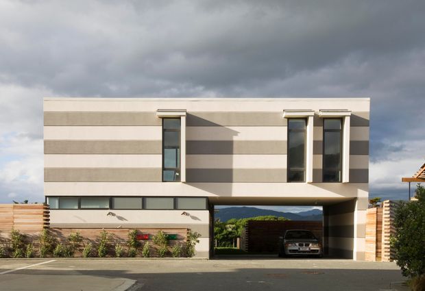 Street-facing exterior of a contemporary two-storey house with a flat roof and strongly horizontal striped facade in pale beige and grey. Narrow vertical windows project from the upper level, while a long horizontal window band, timber fencing, landscaped planting, and an integrated ground-floor carport define the lower facade. A vehicle is visible in the carport, with mountains and cloudy sky beyond.