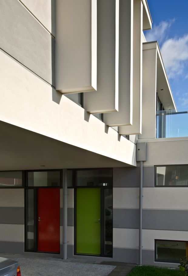 Exterior architectural photograph of a contemporary multi-level building with a light rendered facade, projecting vertical fins, dark horizontal wall bands, large dark-framed glazing, and bold red and lime-green entry doors. A glass-balustraded upper terrace and strong geometric overhangs are visible against a blue sky.