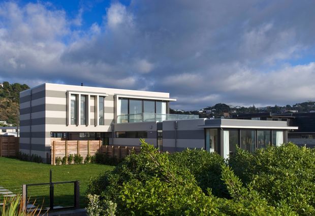 Exterior view of a contemporary multi-level residential house with strong horizontal and rectangular forms, flat roofs, extensive glazing, a projecting upper balcony, and contrasting light and dark facade finishes. The home sits in a landscaped suburban setting with hedges and lawn, with neighbouring homes and a wooded hillside visible behind it.