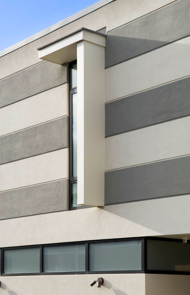 Close-up exterior view of a contemporary building facade featuring horizontal bands of contrasting light and dark grey textured render, a projecting box-like window surround, narrow vertical glazing, and dark-framed horizontal windows at lower level. The composition is geometric and minimalist, with a flat roofline and clean rectilinear detailing against a clear blue sky.