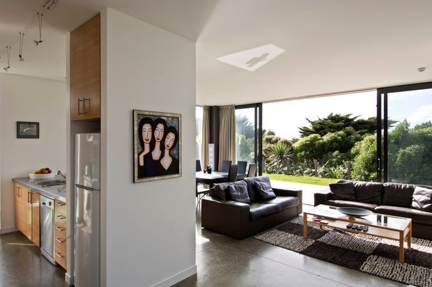 Open-plan contemporary home interior showing a compact kitchen on the left and a bright living room beyond, separated by a white structural wall. The living area has dark leather sofas, a low timber coffee table, a textured dark rug, full-height corner glazing and views to a landscaped garden. The kitchen features timber cabinetry, stainless-steel appliances and a light benchtop. A simple linear pendant installation and recessed skylight add architectural interest.