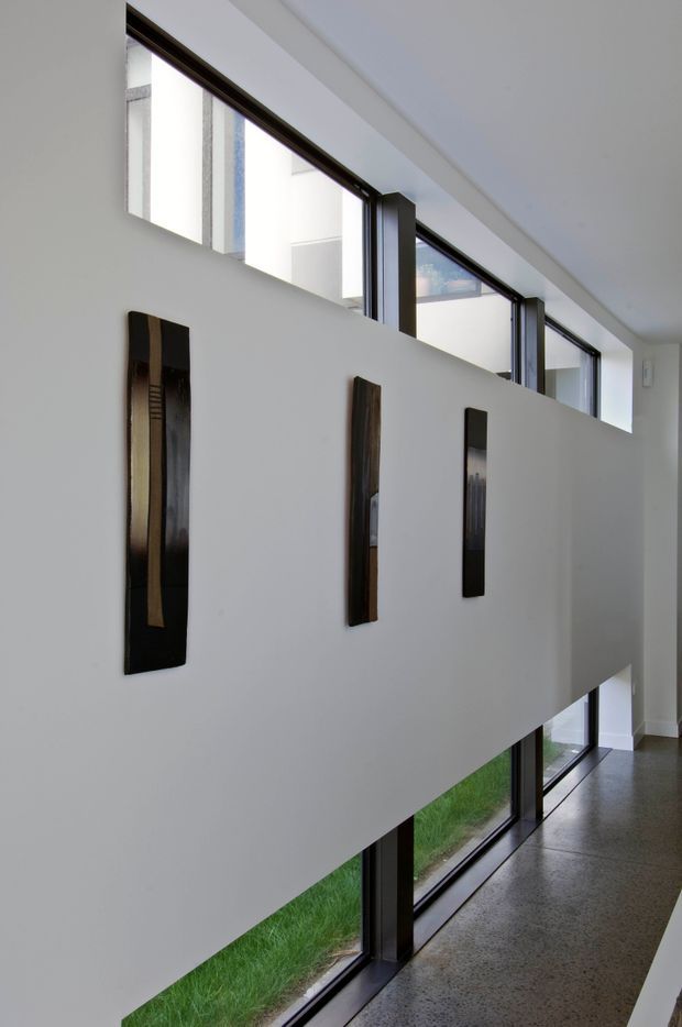 A minimalist interior hallway or landing with a tall white partition wall displaying three narrow dark vertical art panels. Continuous clerestory and floor-to-ceiling glazing with dark frames brings in abundant daylight, while a polished grey concrete or terrazzo-like floor runs alongside the windows. The space has clean geometric lines and a contemporary architectural character.