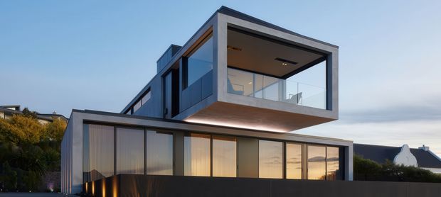 Exterior view of a contemporary multi-level residence at dusk, featuring crisp rectilinear volumes, extensive floor-to-ceiling glazing, a projecting upper-level box with a covered balcony or glazed terrace, dark metal cladding, and warm interior lighting. The home sits behind a dark boundary fence with landscaped surroundings.