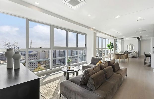 Bright, spacious high-rise apartment living area with floor-to-ceiling windows overlooking dense urban buildings. The open-plan room includes a large taupe sectional sofa, round coffee table, dining area, black kitchen or console joinery, neutral flooring, recessed lighting, and a mirrored wall feature. The interior has a clean, contemporary minimalist feel with abundant daylight.