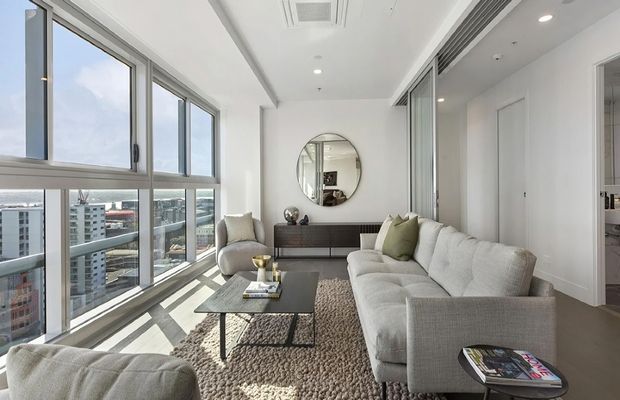 Bright contemporary apartment living room with floor-to-ceiling glazing, expansive city views, a large light-grey sectional sofa, textured area rug, black coffee table, sculptural armchairs, and a slim built-in console beneath a round mirror. The space has a clean white palette, recessed lighting, and a glazed sliding partition or doorway along the right side.