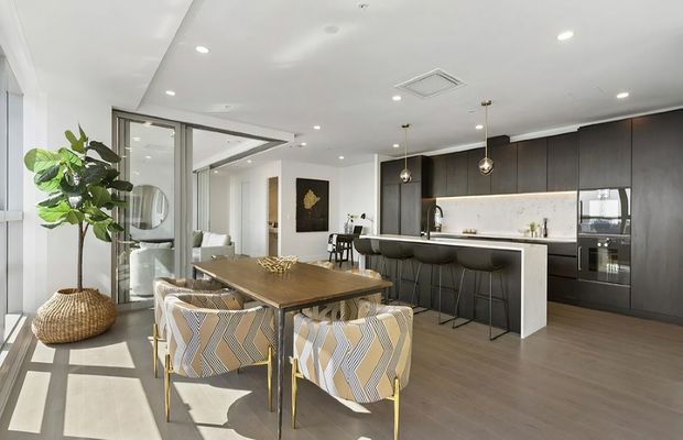 Bright open-plan contemporary interior featuring a dining area and kitchen. A timber dining table with patterned upholstered chairs sits beside full-height glazing, while the kitchen has dark flat-panel cabinetry, a white island and benchtop, integrated appliances, a tiled or stone-look splashback, and pendant lighting. Light wood-look flooring and white walls create an airy, refined setting.