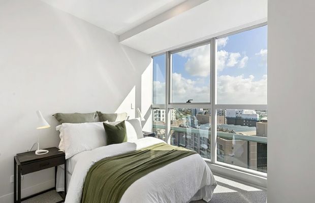 Bright modern bedroom with a neatly made bed, white bedding, an olive-green throw and cushion, a small dark bedside table, and a slim floor lamp. Large floor-to-ceiling windows provide expansive urban views and abundant natural light. The room has white walls, a simple recessed ceiling detail, and grey carpet flooring.