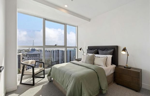 Bright contemporary bedroom in a high-rise apartment, featuring floor-to-ceiling corner glazing with city views, a dark upholstered bedhead, pale green bedding, a timber bedside cabinet, a lounge chair, and minimalist bedside lighting. The room has light carpet, white walls, and a clean, airy finish.