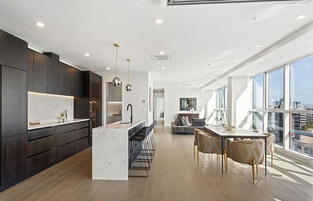 Bright open-plan apartment interior with a contemporary kitchen, large white stone island, dark timber cabinetry, dining area, living room, and expansive floor-to-ceiling windows overlooking an urban setting. The space has light timber flooring, white walls and ceiling, modern pendant lights, and abundant natural light.