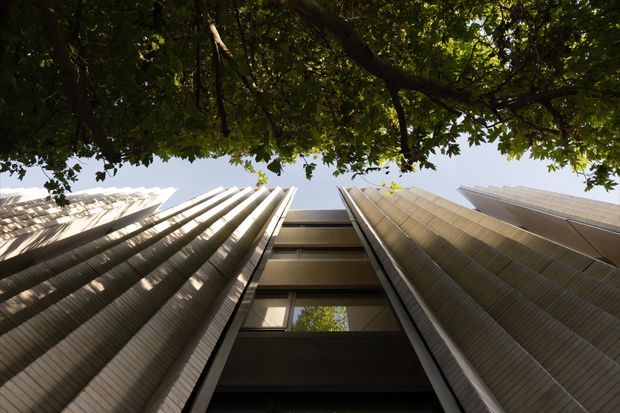 Low-angle architectural photograph looking upward between tall, closely spaced modern building facades. The façades feature strong vertical ribbed or louvred elements in light grey and warm metallic tones, with dark recessed glazing and projecting horizontal slabs. A leafy tree canopy frames the top of the view, creating a contrast between the urban architecture and greenery.