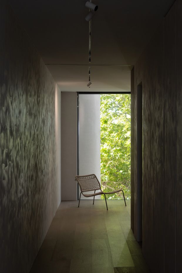 A dark, narrow interior passage or landing with textured dark walls, a pale ceiling with exposed track-style spotlights, and wide timber floorboards. At the far end, a woven lounge chair sits beside a tall opening overlooking dense green foliage, creating a strong contrast between the moody corridor and the bright natural view.