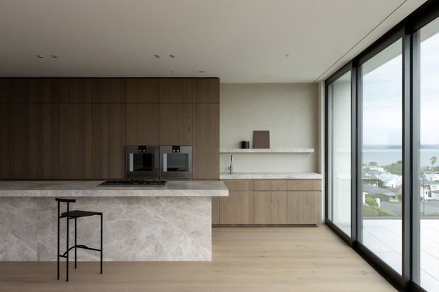 A refined contemporary kitchen with full-height dark timber cabinetry, integrated black appliances, a large pale stone island with waterfall sides, and matching stone splashback and shelving. Light timber flooring and expansive black-framed floor-to-ceiling glazing create an airy interior with coastal views beyond. A minimal black bar stool sits at the island.