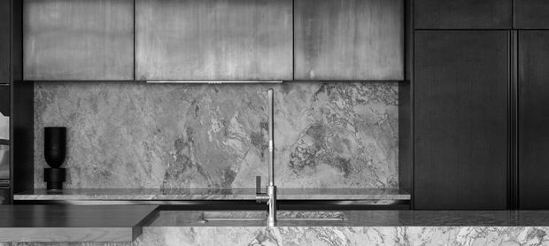Monochrome close-up of a refined contemporary kitchen, featuring dark timber cabinetry, a veined stone splashback and benchtop, brushed metal upper cabinets, and a central stainless-steel tap and sink. The composition focuses on the fixed joinery and material contrast.