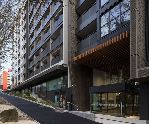 Street-level view of a contemporary multi-storey urban building with a dark, finely patterned facade, extensive glazing, projecting balconies, and a prominent recessed entrance beneath a warm timber-look slatted canopy. The building fronts a landscaped sloping pedestrian approach and appears to include commercial or lobby space at ground level.
