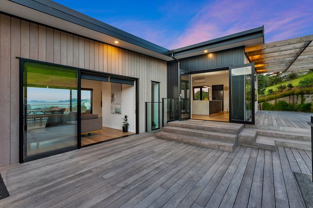 Contemporary single-storey home with a low-pitched roof, dark metal cladding and extensive glazing opening onto a broad weathered timber deck. The stepped deck connects several indoor-outdoor living spaces, with a kitchen visible through the central opening and a living room visible at left. A landscaped sloping garden rises behind a partially covered deck area, while the distant water view is visible through the glazing.