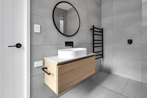 Contemporary bathroom featuring large-format grey wall and floor tiles, a floating timber vanity with a white vessel basin, black wall-mounted tapware, a round black-framed mirror, and a black heated towel rail. The space has a clean, minimalist fit-out with strong monochrome and warm timber contrast.