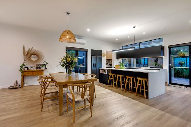 A bright, open-plan contemporary kitchen and dining area. The room features a large light-wood dining table with timber chairs, a substantial dark island with a pale stone benchtop and integrated breakfast bar, black-framed glazing and doors, light timber flooring, white walls, recessed lighting, and woven pendant lights. A timber console and decorative mirror sit along the dining wall.