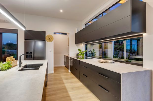 A contemporary kitchen with a large pale stone island, integrated sink and dark tapware, extensive dark matte cabinetry, timber flooring, stainless-steel refrigerator, and a long horizontal window above the benchtop. Under-cabinet lighting and recessed ceiling lights create a clean, bright interior.
