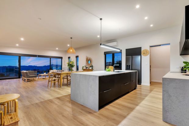 Open-plan contemporary kitchen with a large central island featuring a light grey stone-look end panel and dark cabinetry, opening into a dining and living area with extensive glazing and views beyond. The room has light timber flooring, white walls and ceiling, pendant and recessed lighting, and a restrained neutral palette.