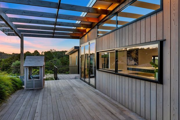A contemporary timber-clad building viewed from a broad outdoor deck or terrace at dusk. The space features vertical grey timber wall cladding, extensive black-framed glazing, a covered pergola with exposed timber beams and translucent roof panels, and timber decking. A small enclosed structure sits at the edge of the deck, with landscaped greenery and a wooded landscape beyond. Warm interior lighting and a visible interior kitchen or living area create an inviting connection between inside and out.