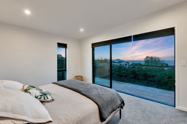 A contemporary bedroom with a low-profile bed, neutral layered bedding, recessed ceiling lights, and pale carpet. Full-height black-framed sliding glazing opens to a timber deck and glass balustrade, overlooking a coastal or waterfront setting at dusk.