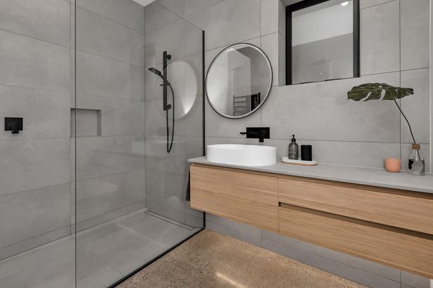 Contemporary bathroom with a walk-in glass shower, large-format grey tiled walls and floor, a floating light timber vanity with a white vessel basin, matte black tapware and hardware, round mirror, and minimal styling with a potted plant.