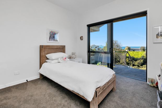 Bright, simply furnished bedroom with a timber bed frame and headboard, white bedding, carpeted floor, white walls, and large black-framed sliding glass doors opening to an outdoor balcony or terrace with coastal views. The room has a clean, contemporary feel and receives abundant natural light.