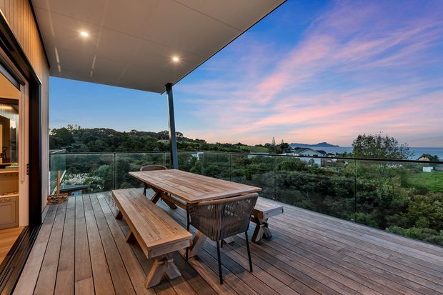 A spacious timber-clad outdoor deck and covered terrace overlooking a lush coastal landscape and water. The deck features a long wooden dining table with benches and chairs, frameless glass balustrading, recessed ceiling lights, and direct connection to the interior through glazed doors. The setting is captured at dusk with a pink and blue sky, creating a relaxed, scenic outdoor entertaining area.