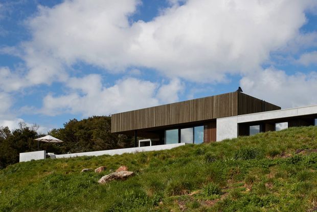 Exterior view of a contemporary single-storey house set into a grassy, sloping landscape. The building has a low horizontal profile, a flat roof, dark vertical timber cladding forming a projecting upper fascia, white rendered masonry walls, and large dark-framed windows. A terrace and outdoor umbrella are visible to the left, with trees behind the house and a partly cloudy sky.
