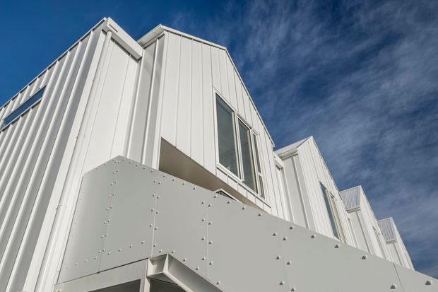 Low-angle exterior view of a contemporary multi-storey building with crisp white vertically ribbed cladding, projecting geometric volumes, tall windows, and a prominent pale metal or panelled overhang with exposed fasteners. Blue sky forms the background.