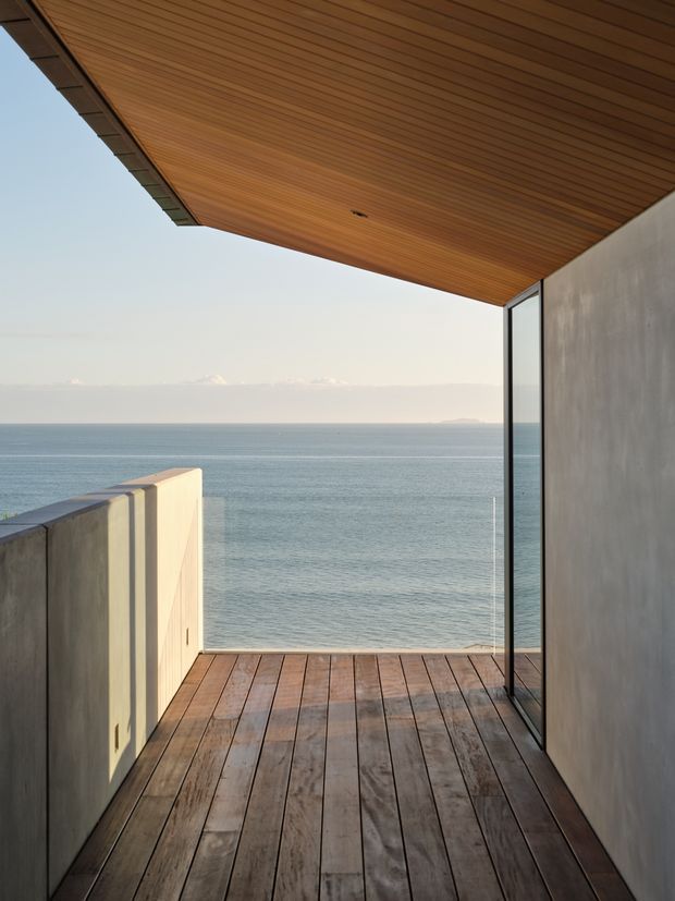 A minimalist coastal balcony or terrace overlooking the ocean. The space features a warm timber-lined soffit, timber decking, smooth concrete side walls, and full-height glazed doors or windows. The clean architectural framing creates an open, serene outlook toward the sea and distant horizon.
