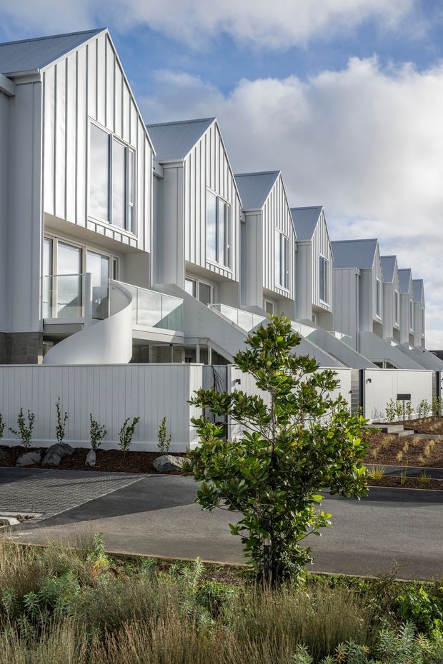 Exterior architectural photograph of a contemporary row of attached multi-storey townhouses. The buildings feature repeated steep gable forms, pale vertical weatherboard-style cladding, grey metal roofing, large glazed windows, and curved white balcony or stair elements with glass balustrades. A landscaped streetscape with young trees, grasses, paving, and dark asphalt occupies the foreground under a partly cloudy sky.