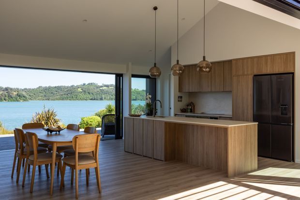 A contemporary open-plan kitchen and dining area with warm timber cabinetry, a large island, integrated dark appliances, pendant lights, and a timber dining table with chairs. Full-height glazing opens the room toward a sunny waterfront view, while dark timber-toned flooring and strong sunlight create a warm, inviting atmosphere.