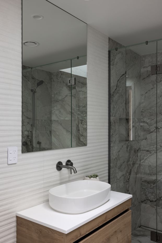 Contemporary bathroom vanity with a white vessel basin, wall-mounted dark metal tapware, timber-look drawer cabinetry and a white benchtop. The walls feature horizontally textured light tiles, while the enclosed shower is lined with dramatic grey marble-look slabs and clear frameless glass. A large mirror reflects the shower area and recessed ceiling lighting.