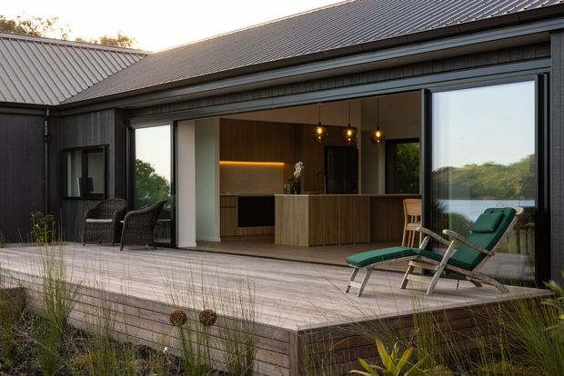 Exterior view of a dark-clad, single-storey pavilion-style home with a pitched metal roof and broad sliding glass openings onto a timber deck. The open interior reveals a compact contemporary kitchen with a timber island, dark cabinetry, warm under-cabinet lighting, and pendant lights. Outdoor lounge chairs and landscaped grasses occupy the foreground, while a calm water view is visible beyond the glazing.