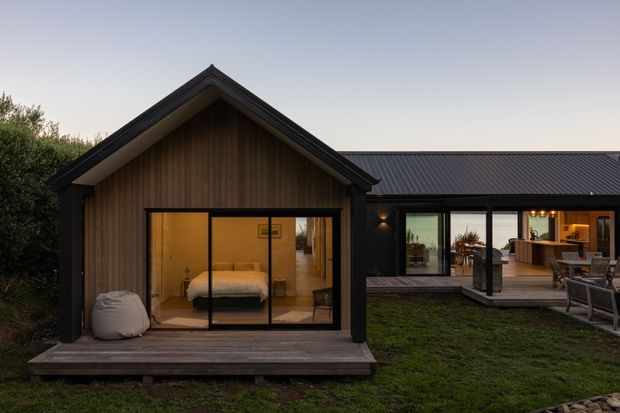 Exterior view of a modern single-storey home at dusk, composed of a compact gabled timber-clad pavilion connected to a longer flat-profile wing with dark standing-seam roofing and extensive black-framed glazing. The foreground pavilion opens onto a timber deck and contains a visible bedroom, while the right-hand wing reveals an illuminated kitchen and dining/living area. The property is surrounded by lawn and planting, with warm interior lighting contrasting against the dark exterior finishes.