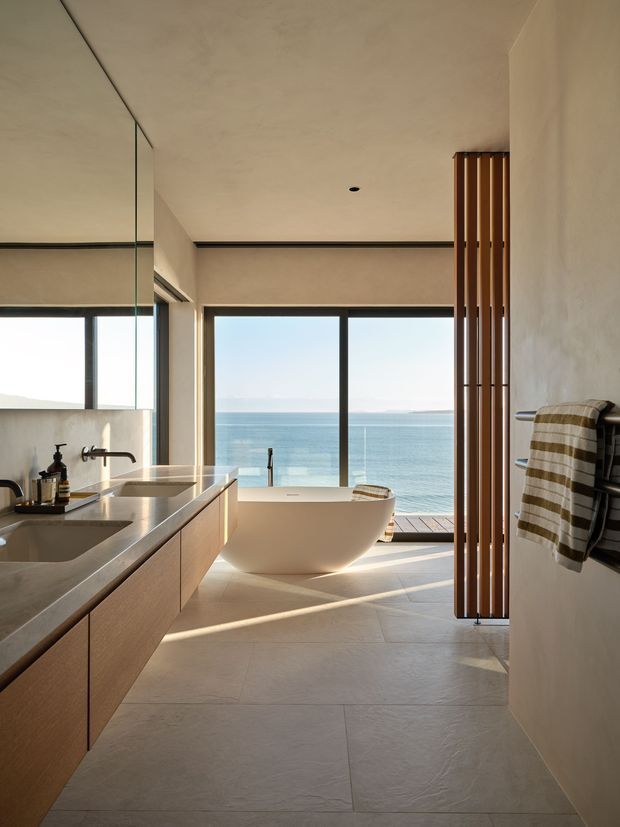 A contemporary bathroom with a freestanding white bathtub positioned beside full-height ocean-facing glazing. The room features a long timber vanity with an integrated pale stone benchtop and basin, large light stone-look floor tiles, a frameless mirror, black tapware, and a vertical timber slatted privacy screen. Warm sunlight creates a calm, refined coastal atmosphere.