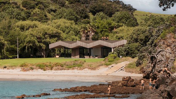 A low-profile contemporary house set directly beside a sandy beach and rocky shoreline, surrounded by dense native vegetation and rolling green hills. The building has a distinctive folded or winged roofline, extensive dark-framed glazing, and timber-toned exterior walls, creating a strong connection between the interior and coastal landscape.