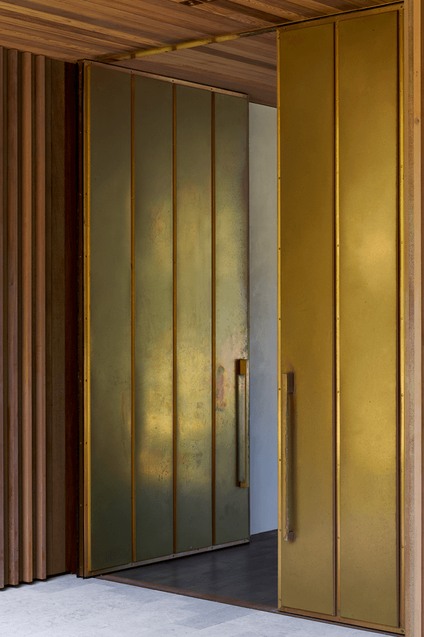 Close-up architectural view of tall pivot or hinged doors with aged brass-toned metal panels and vertical raised strips. The doors open into a dark interior, framed by warm vertical timber wall battens and a timber-lined ceiling. A pale smooth floor and raw, understated doorway surround complete the industrial, tactile composition.