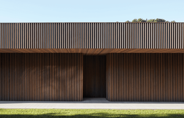 A minimalist, single-storey building facade with a strong horizontal flat roofline and continuous vertical timber-look slatted cladding. The dark ribbed facade wraps around a recessed central entry, with a timber-lined soffit above a pale paved walkway and lawn. Clear blue sky and a small amount of planting are visible above the roof.