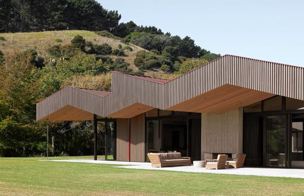 A low-slung contemporary residence set against a densely planted hillside. The building has sharply folded, corrugated vertical-clad roof forms with deep timber-lined soffits, extensive dark-framed glazing, and a pale textured wall surface. A broad lawn and paved terrace sit in front, with outdoor seating beneath the covered overhangs.