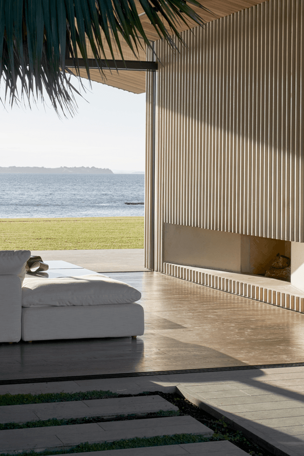 A refined open-sided coastal living space opening directly onto a lawn and ocean view. The room features a large white sectional sofa, polished warm-toned concrete flooring, a timber-lined ceiling, and a striking full-height wall of closely spaced vertical timber slats incorporating a recessed fireplace. Broad openings and deep overhangs create strong sunlight and shadow patterns, while large stepping stones connect the interior to the exterior.
