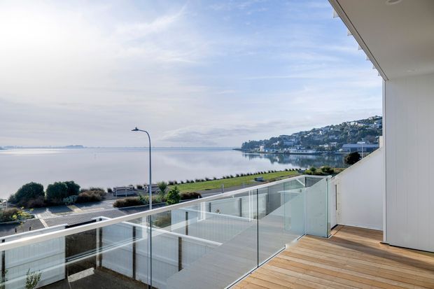 A bright modern balcony or terrace overlooking a calm waterfront bay. The space features warm timber decking, frameless glass balustrades, white exterior walls and soffits, and clean contemporary architectural lines. A landscaped waterfront promenade and hillside settlement are visible beyond the terrace.
