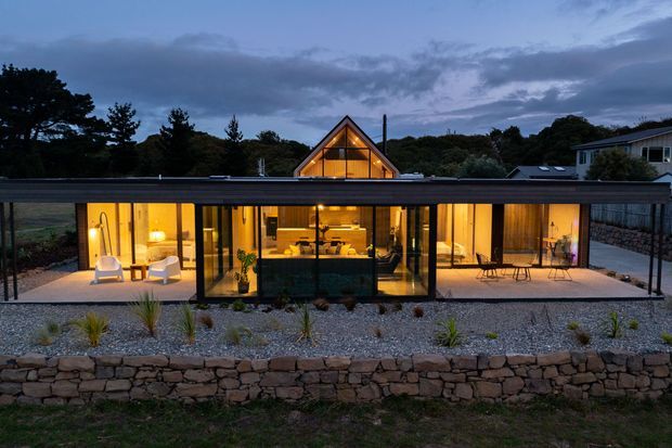 Wide dusk exterior view of a contemporary single-storey home with a long, low flat-roofed glazed pavilion in the foreground and a taller illuminated gable-roofed volume behind it. Extensive floor-to-ceiling glazing reveals warm-lit living spaces and a central kitchen, while a gravel garden, stone retaining wall, deck or terrace, and surrounding trees frame the building.