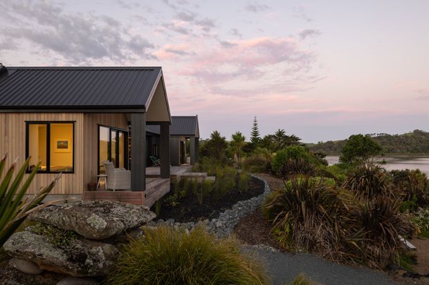 Exterior view of a low-rise contemporary dwelling beside a calm body of water at dusk. The building has dark corrugated metal gable roofs, vertical timber-like wall cladding, large glazed openings, and a small timber deck. Native-style landscaping with rocks, grasses, shrubs, and sculptural plants surrounds a gravel garden path.