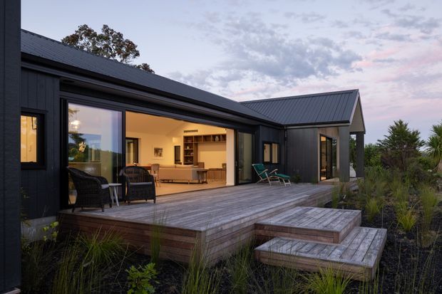 Exterior view of a contemporary single-storey dark-clad home at dusk, with intersecting pitched roof forms, large glazed openings, and a broad timber deck with stepped platforms. The open living area and built-in shelving are visible through the glazing, while native-style planting surrounds the deck.