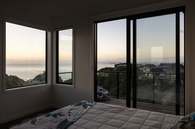 A bedroom viewed from beside the bed, with expansive black-framed windows and glazed sliding doors opening to a balcony. The room overlooks a coastal landscape and ocean at dusk, with neutral walls, a quilted bedspread, and a calm, subdued atmosphere.
